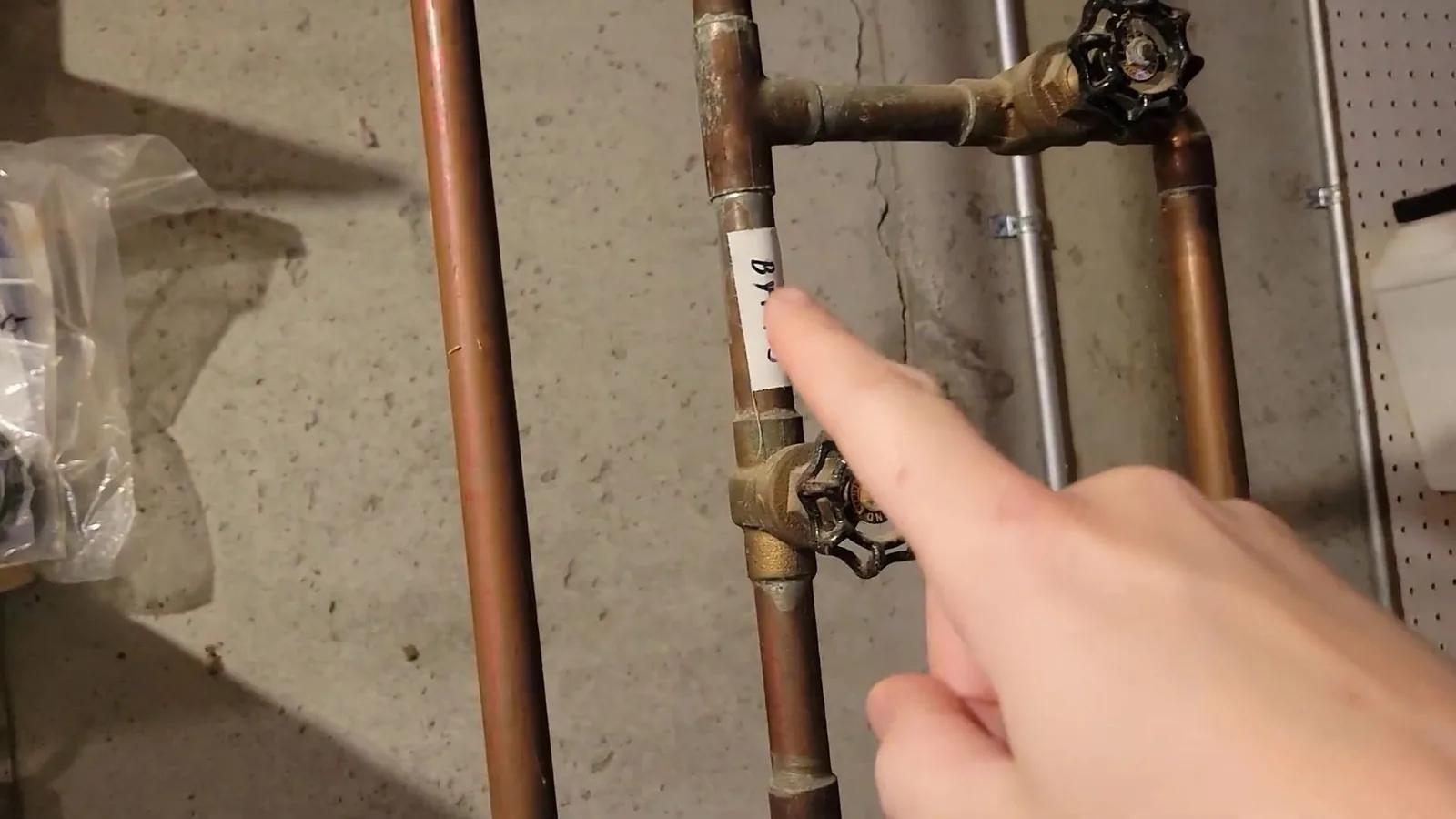 A finger pointing at a brass shutoff valve on copper water pipes along a basement wall